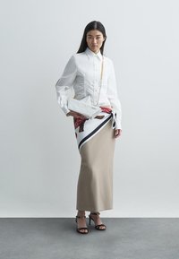 Biała koszula z guzikami i bufkowymi rękawami, beżowa spódnica z czarnymi i czerwonymi akcentami w wzory, trzymająca białą torebkę kopertówkę. Czarne sandały na paski.