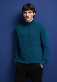 Pull en col roulé côtelé teal à manches longues, avec une texture de rayures subtiles. Associé à un pantalon noir, sur fond bleu.