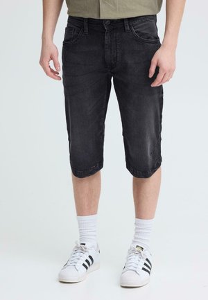 Man wearing black knee-length denim shorts, white crew socks, and white Adidas sneakers with black stripes, standing against a plain background.