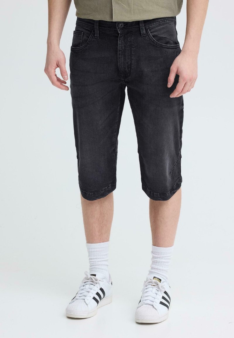 Άνδρας που φοράει μαύρα τζιν σορτς μέχρι το γόνατο, λευκές κάλτσες μέχρι τον αστράγαλο και λευκά αθλητικά παπούτσια Adidas με μαύρες ρίγες, στέκεται μπροστά σε απλό φόντο.