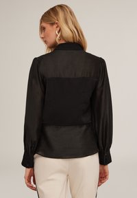 Blusa nera con un design colletto, maniche lunghe a sbuffo e polsini con bottoni. Tessuto liscio, leggermente trasparente, con schiena aderente.