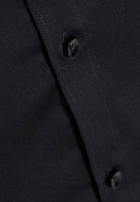 Gros plan sur un tissu noir avec deux boutons noirs cousus sur un vêtement, montrant les coutures et les boutonnières sur un matériau lisse.