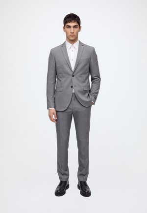 Joven de pie vistiendo un traje gris de dos botones con pantalones a juego, camisa blanca y zapatos de vestir negros, sobre un fondo liso y claro.
