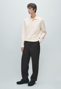 Kremowa koszulka polo z długim rękawem i plisą na trzy guziki, zestawiona z ciemnymi, szerokimi spodniami i czarnymi zamszowymi loafersami. Prosty i elegancki design.