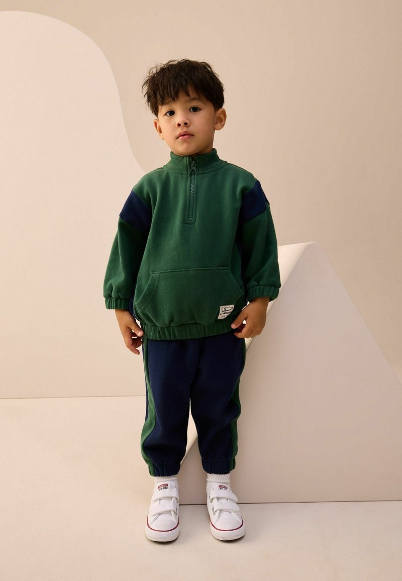 Survêtement vert et bleu marine en tissu doux, doté d'un col zippé à mi-hauteur, de poignets élastiques et d'une poche avant. Porté avec des baskets blanches.
