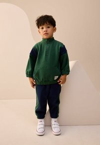 Grøn og marineblå træningsdragt lavet af blødt stof, med en halv-lynlås krave, elastiske ærmer og en frontlomme. Båret med hvide sneakers.