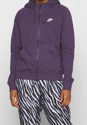 Personne portant un sweat à capuche zippé Nike violet avec des poches avant et un pantalon à imprimé zèbre noir et blanc, debout devant un fond uni.