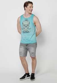 Turkos linne med ett dödskallemönster, kombinerat med grå denimshorts med fransar och revor, kompletterat med svarta sneakers.