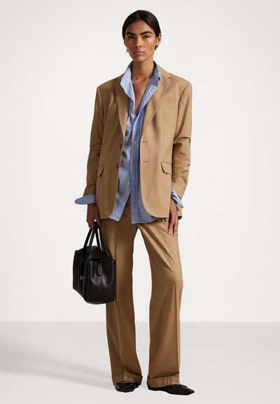 Femme portant un costume beige avec un pantalon large, une chemise bleu clair, tenant un sac à main noir, se tenant devant un fond blanc.