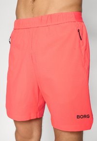 Calções de cor coral brilhante, com uma superfície texturizada, faixa elástica na cintura, bolsos laterais com fechos éclair e um detalhe de logótipo preto.