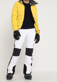 Doudoune jaune avec doublure grise, pull navy, pantalon de ski blanc avec accents noirs, gants noirs et chaussures de ski.