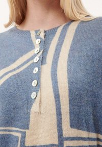 Gros plan sur un pull tricoté bleu et beige avec des rayures verticales et diagonales, comprenant une rangée de sept boutons blancs près de l'encolure.