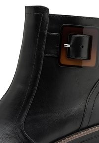 Bottine en cuir noir avec une boucle en écaille de tortue carrée, texture lisse et coutures contrastantes claires le long de la semelle.