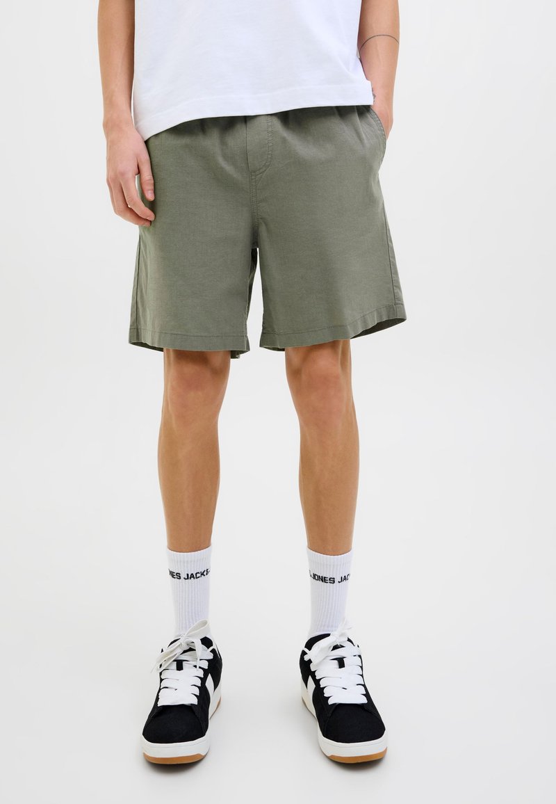 Pantalones cortos verde caqui, material de algodón, ajuste relajado, bolsillo laterales, dobladillo acabado, usados con zapatillas negras y calcetines blancos.