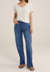 Kvinna iklädd en beige och vit randig V-ringad T-shirt, högmidjade raka blå jeans och tanfärgade sandaletter med öppen tå och spänne.