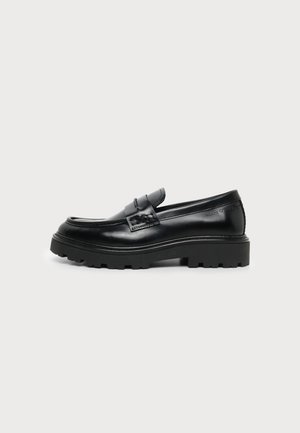 Zwarte leren loafer met dikke profielzool en penny-band detail, getoond vanaf de zijkant op een effen lichte achtergrond.