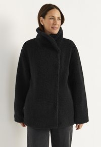 Mustapintainen fleece-takki, jossa on korkea kaulus, rento istuvuus ja tekstuuri. Takissa on etulukko ja sivutaskut.
