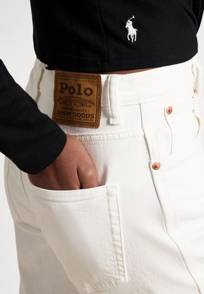 Person trägt weiße Polo Ralph Lauren Jeans mit der Hand in der Gesäßtasche und ein schwarzes Hemd mit weiß besticktem Polospieler-Logo.