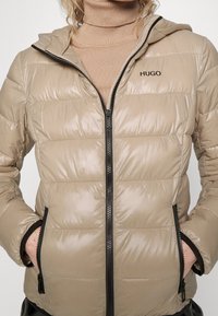 Beżowa kurtka puchowa o połyskującej powierzchni, z kapturem, zamkiem z przodu i czarnymi akcentami. Posiada poziomo pikowane panele oraz boczne kieszenie.