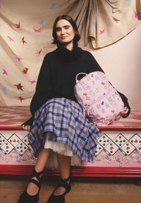 Rózsaszín hátizsák játékos mintákkal, fekete túlméretezett pulóver, kék kockás szoknya és fekete cipő keresztpántokkal.