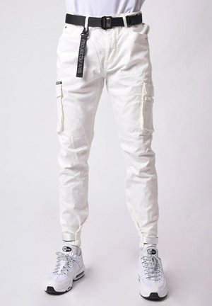 Pantaloni cargo bianchi con multiple tasche, tessuto texturizzato, vestibilità affusolata e una cintura nera con etichetta di marca. Abbinati a sneakers bianche.