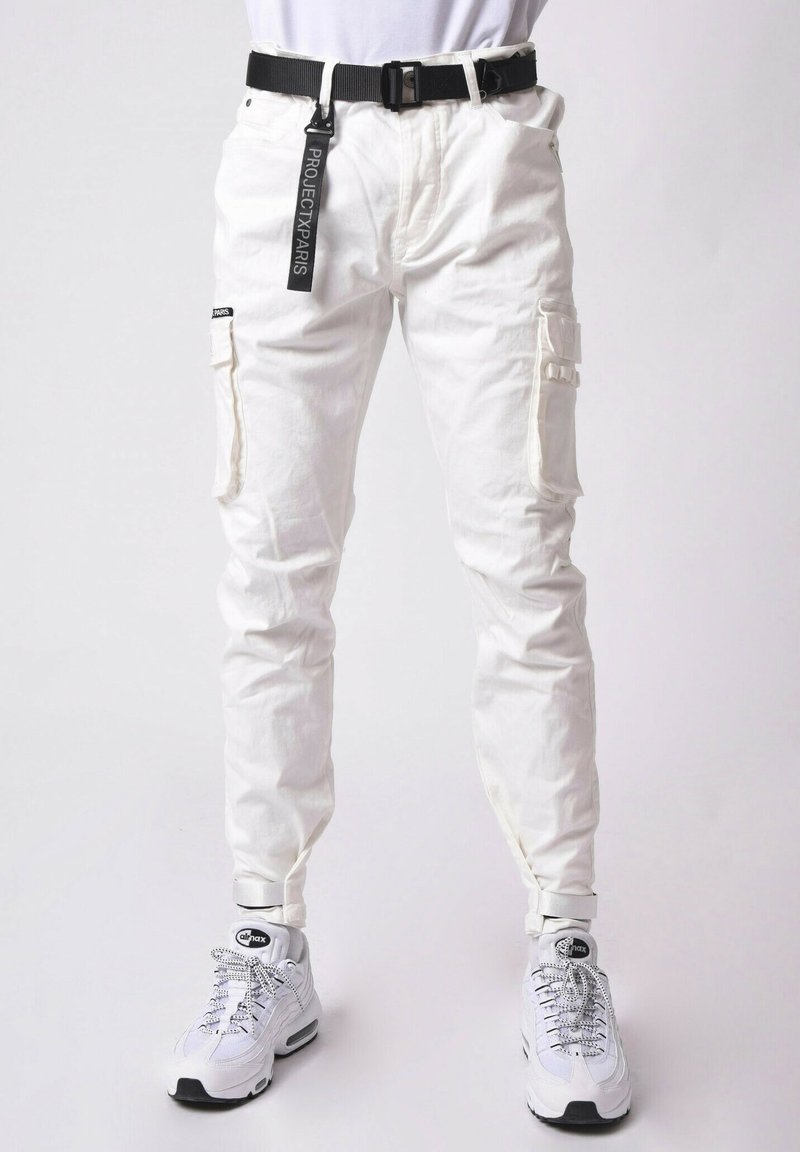 Pantalones cargo blancos con múltiples bolsillos, una textura de tela, corte ajustado y un cinturón negro con una etiqueta de marca. Combinados con zapatillas blancas.