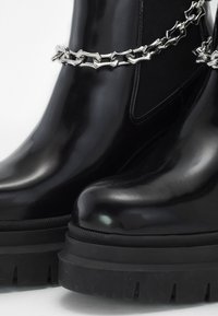 Sorte, blanke ankelstøvler med en kraftig gummisål, der har en sølvkæde-detalje omkring overdelen og elastiske sidepaneler for pasform.