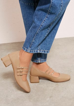 Beige Lackleder-Pumps mit quadratischer Spitze, versehen mit drei dekorativen Riemen und Schnallen. Blockabsatz-Design, getragen zu blauem Denim.