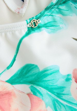 Audimo raštuotą audinį, kuriame pavaizduoti rožiniai rožės ir žalios lapai, su auksiniu užsegimu netoli kaklo, ant balto fono.