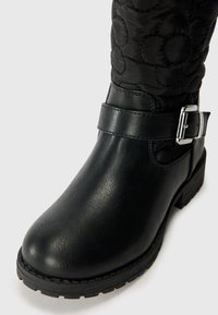 Botín de cuero negro con punta redondeada, acabado mate, caña de tela acolchada con patrones florales y un detalle de hebilla lateral.