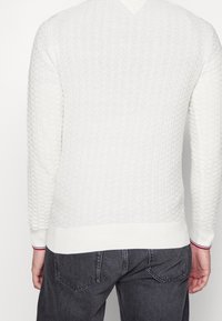 Pull en tricot blanc avec un motif texturé, décolleté en V, poignets et ourlet côtelés ; associé à un jean gris foncé.