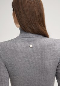 Maglione grigio a collo alto con una decorazione di bottoni sul retro. Il tessuto sembra morbido con una finitura testurizzata.
