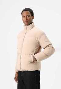 Beige donsjas met hoge kraag, ritssluiting, zijzakken en elastische manchetten. Gladde textuur en doorgestikt ontwerp.