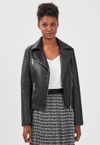 Giacca biker nera in ecopelle con zip argentate, ampie revers e una vestibilità corta, indossata sopra una maglietta bianca e una gonna a fantasia.