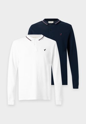 Homme aux cheveux bouclés portant un polo blanc à manches longues, un jean bleu ample et des chaussures marron, debout devant un fond blanc uni.