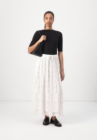 Zwarte getailleerde top, witte maxi rok van kant met patroon en strik taille, gecombineerd met zwarte loafers en een kleine zwarte handtas.
