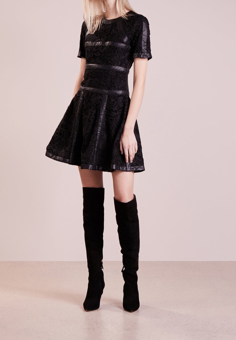 Femme portant une courte robe noire en dentelle avec des bordures en cuir, associée à des bottes en daim noires montantes jusqu’aux genoux, debout sur un sol clair avec un fond beige.