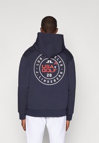 Marineblå hettegenser med et stort sirkulært grafisk design på baksiden som viser "USA GOLF" og "28." Laget av bomullsmateriale med en myk tekstur og kengurulomme.