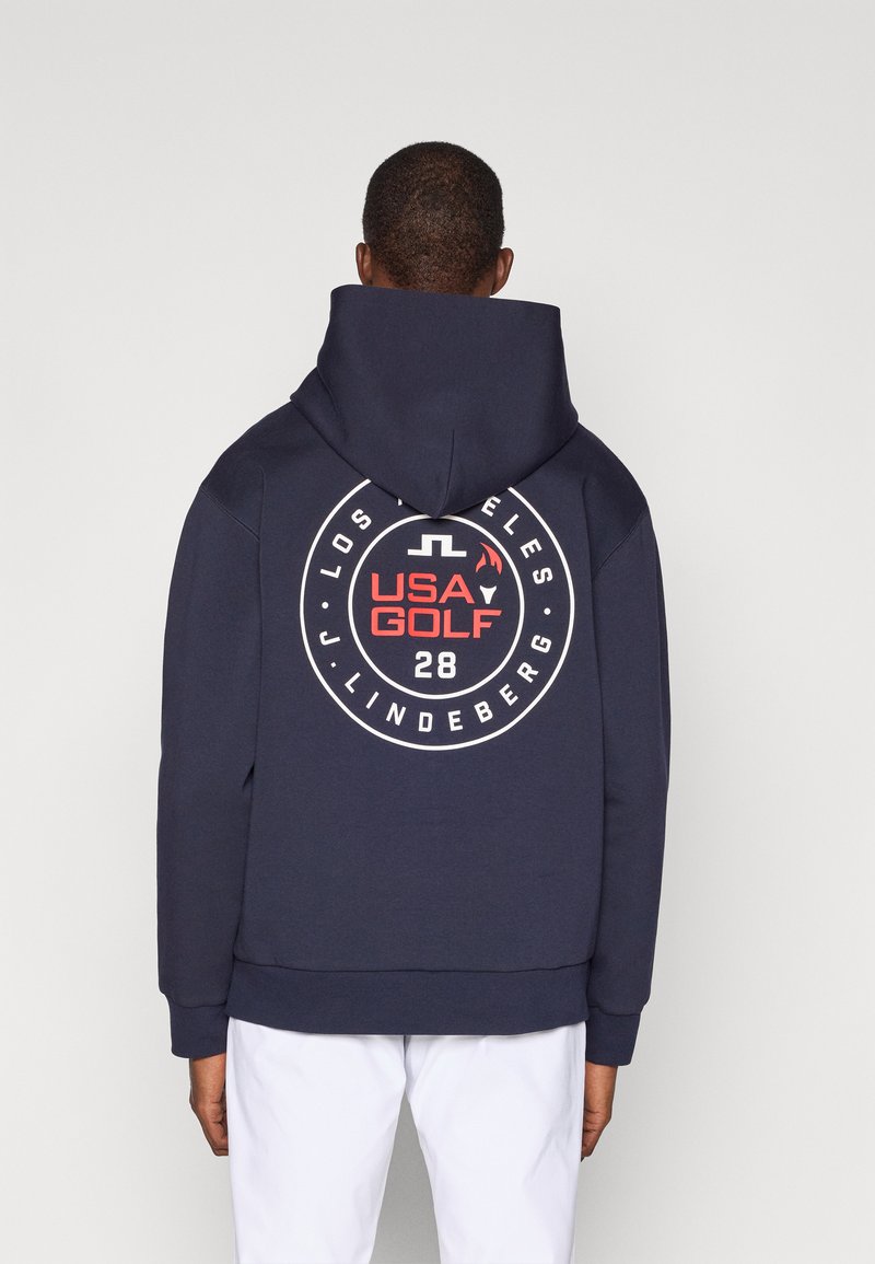 Marineblå hettegenser med et stort sirkulært grafisk design på baksiden som viser "USA GOLF" og "28." Laget av bomullsmateriale med en myk tekstur og kengurulomme.