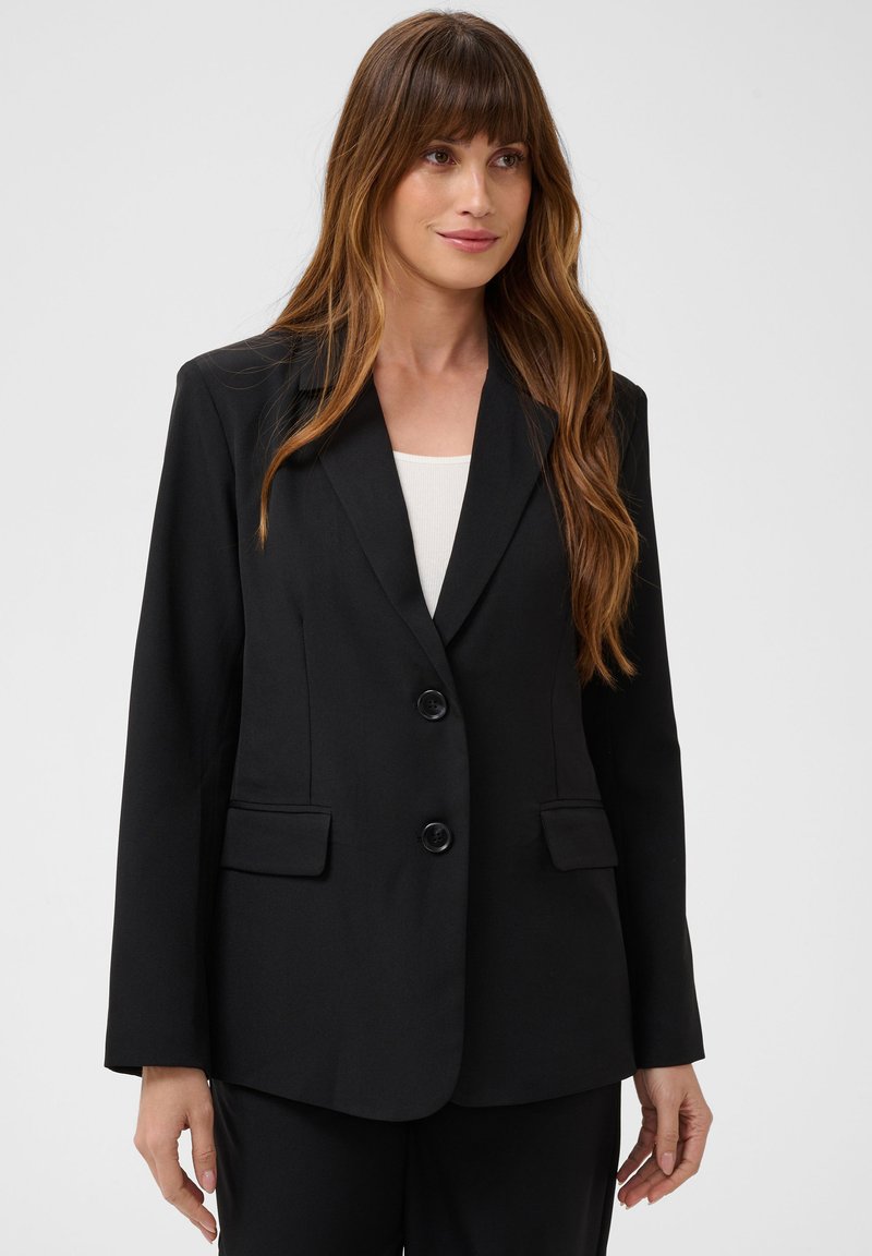Femme aux longs cheveux bruns portant un blazer noir à deux boutons par-dessus un top blanc, debout devant un fond clair uni.