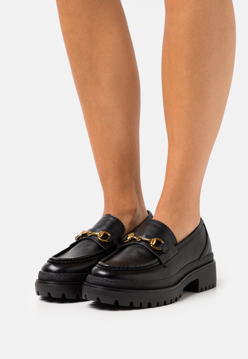 Mocassins chunky en cuir noir avec détail en métal doré portés sur des jambes nues sur un fond uni.