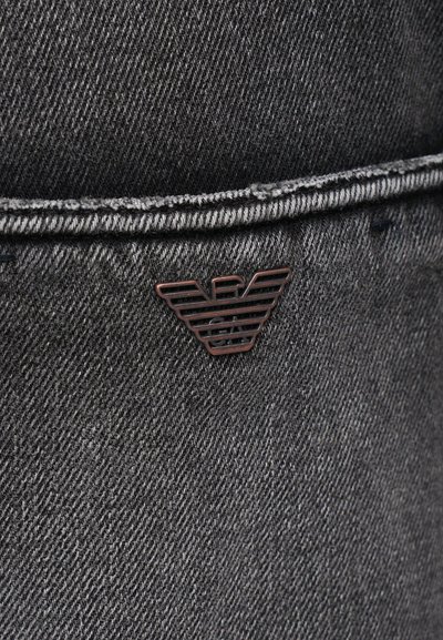 Tmavohnedá džínsovina s hladkou textúrou, so bronzovým emblémom stylizovaných krídel pri šve vrecka.