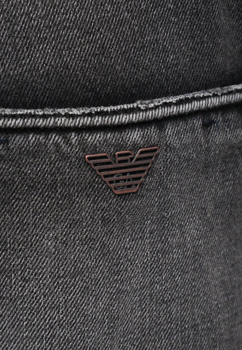 Tissu en denim gris foncé avec une texture lisse, présentant un emblème en bronze de ailes stylisées près de la couture de la poche.