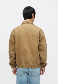 Barna farmerdzseki laza szabással, gumírozott szegéllyel és gallérral. Hátulról látható, világoskék farmernadrág fölött viselve.