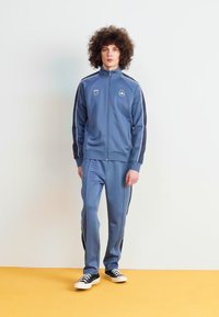 Chándal azul con mangas largas y chaqueta con cremallera, presentado con rayas blancas a lo largo de los costados. Combinado con zapatillas negras.