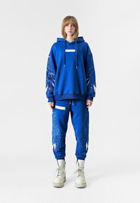 Sudadera azul y pantalones de chándal con intrincados bordados de alas en azul y blanco, con cordones y detalles en blanco. Combinados con botas beige.