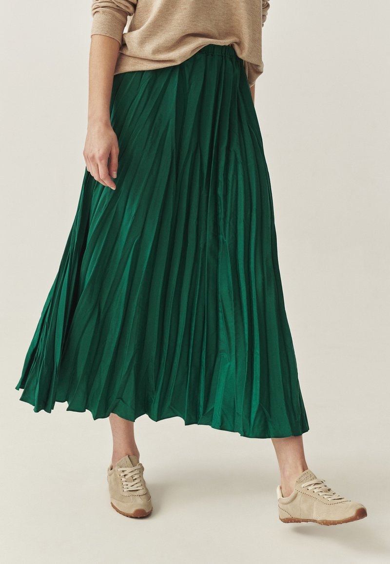 Falda midi plisada verde en tejido ligero, combinada con zapatillas beige. La falda tiene una cinturilla elástica y una textura fluida.