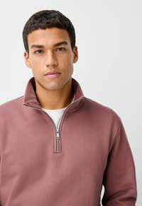 Półzapinany pulower w kolorze przykurzonego różu, wykonany z miękkiej mieszanki bawełny z teksturowanym materiałem. Posiada stojący kołnierz i srebrny detal zamka. Gładka wewnętrzna podszewka.