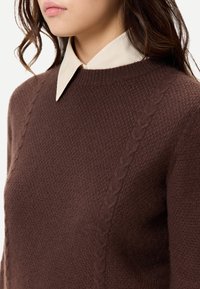 Kvinna som bär en brun texturerad stickad tröja med kabelmönster och en krämfärgad skjorta med krage under, visad från hals till bröst.