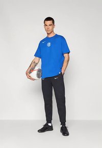 T-shirt bleu à manches courtes avec logo, pantalon noir fuselé et chaussures de football noires. Tenant un ballon de football noir et blanc à motif.